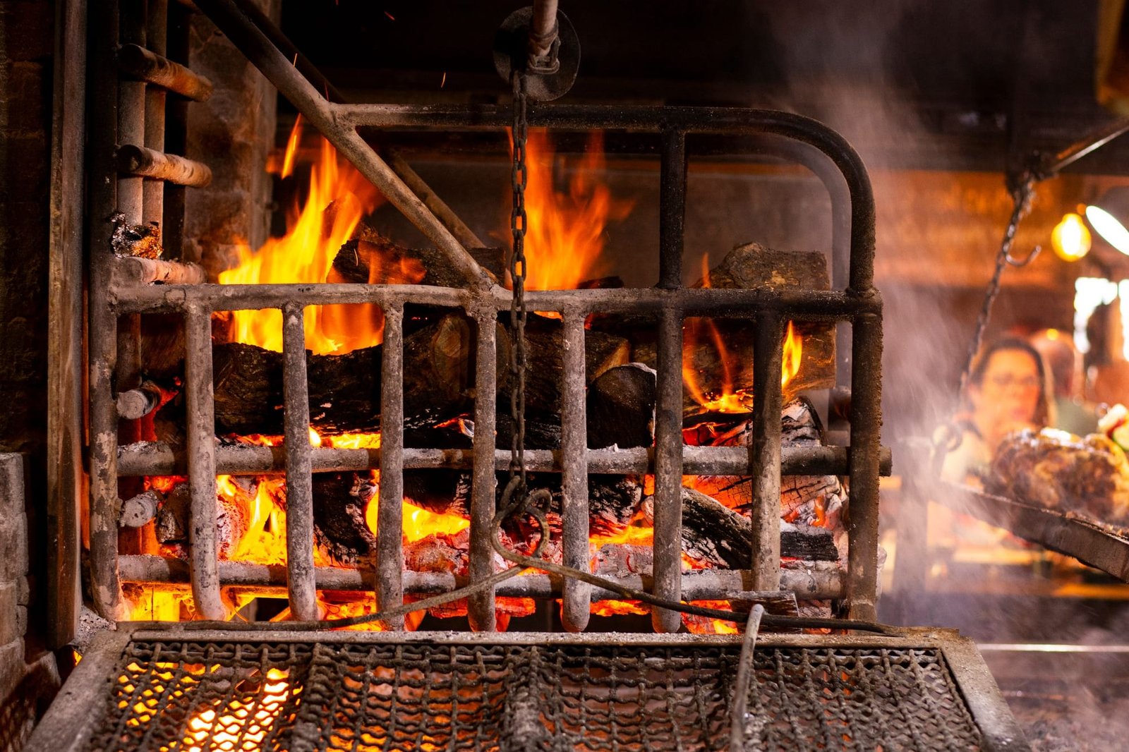 Mesquite and oak burning in the iron fire basket, a cook working behind it