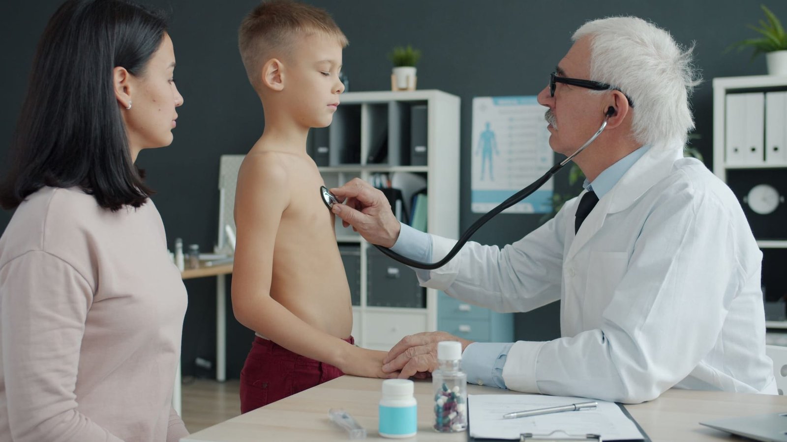 Dr. Bell listens to a boy's chest with a stethoscope while the boy's mother stands beside him in an exam room