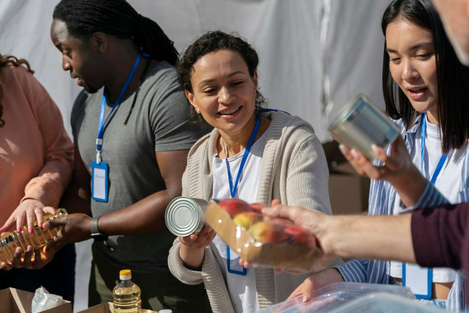 Volunteers with lanyards at an outdoor table, smiling as they pass cans, cooking oil and a tray of peaches into boxes