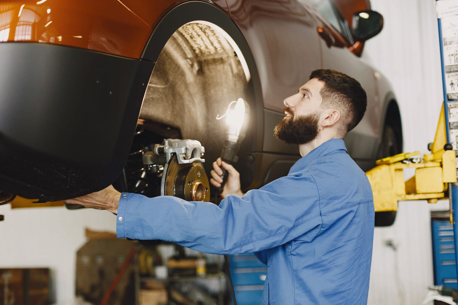 An orange crossover up on the lift with its front wheel off while a mechanic checks the brake rotor with a work light