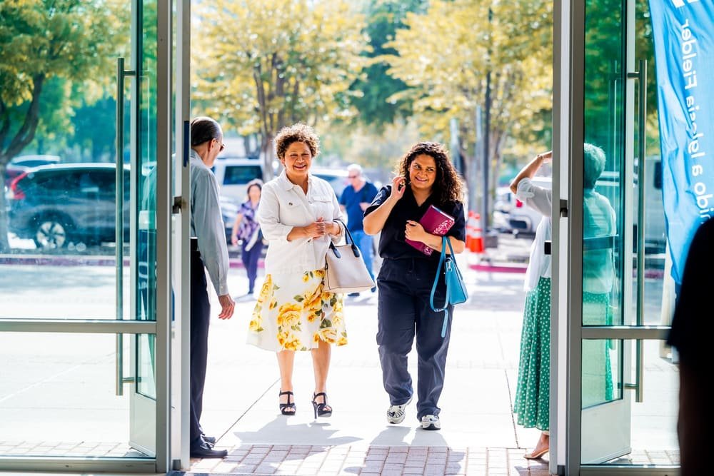 Two women walking in through the open front doors on a sunny morning while a greeter holds the door