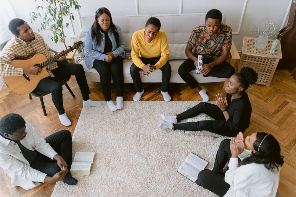 A small group sitting on the floor and couch of a living room with a guitar and open Bibles