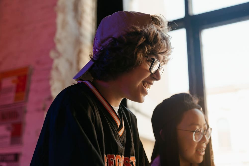 Two teenagers laughing together by a bright window on a Wednesday night