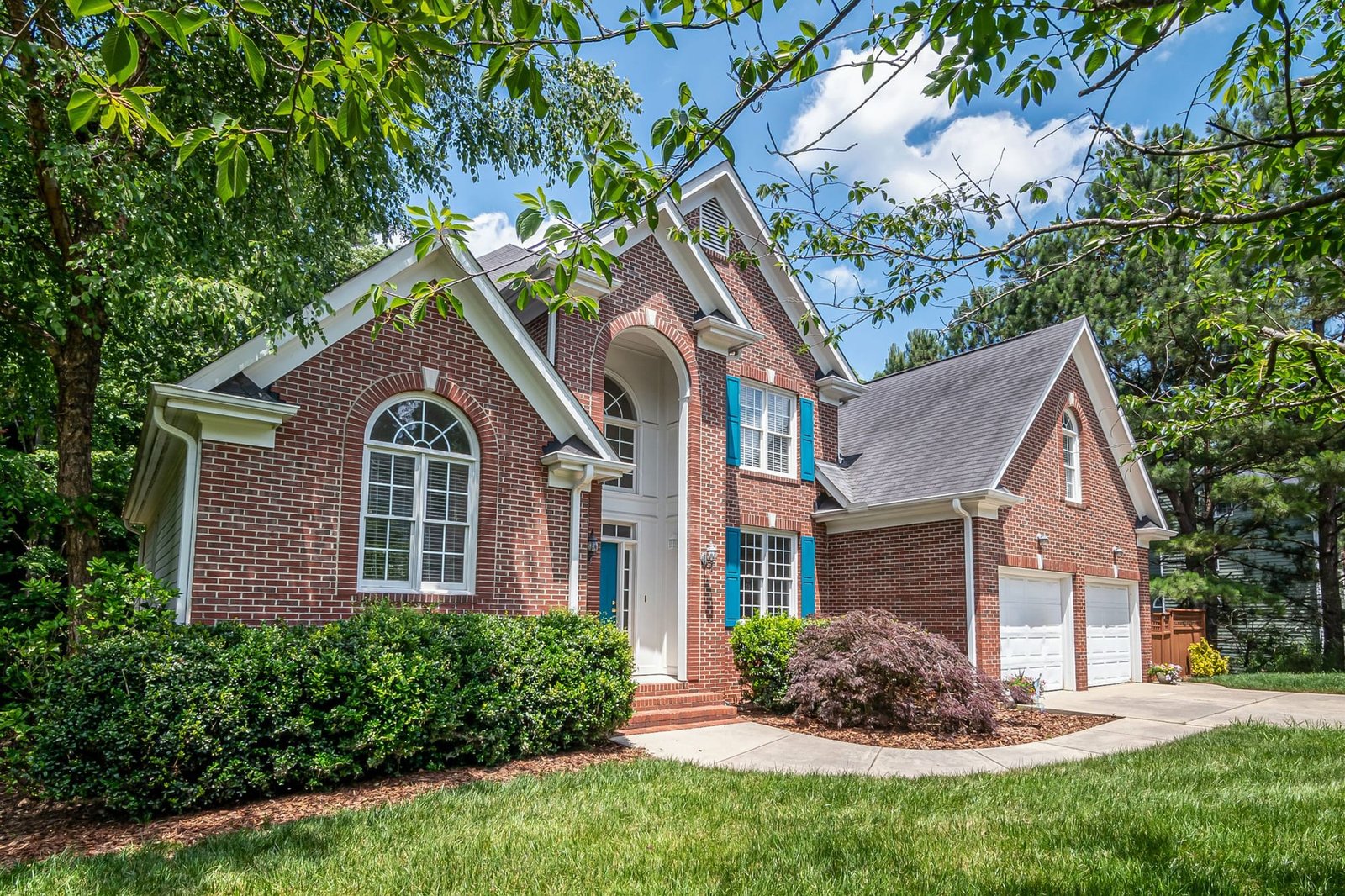 A red brick two story house with blue shutters, a row of round boxwoods in a mulched bed, a red Japanese maple by the door, a curving walk and a fresh cut lawn