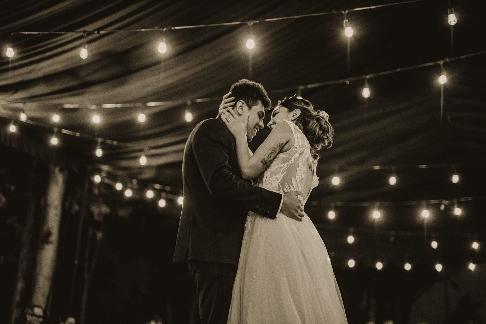 A couple laughing forehead to forehead during their first dance under string lights