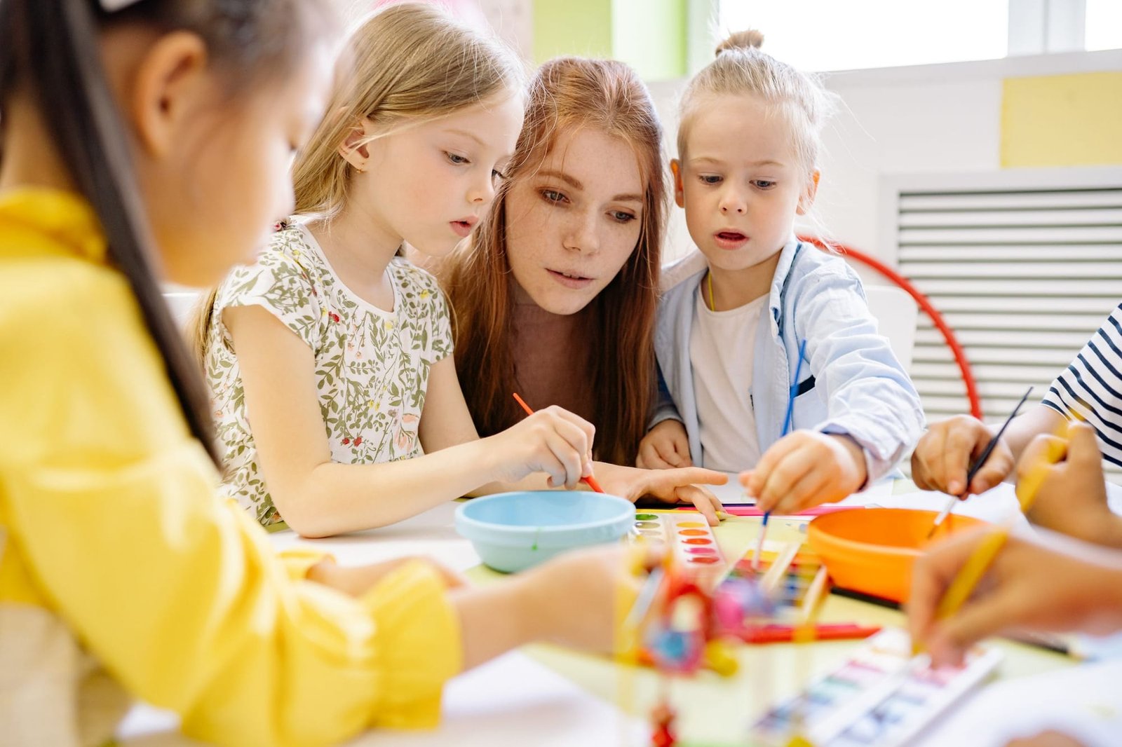 A teacher painting with watercolors alongside three preschoolers at a table