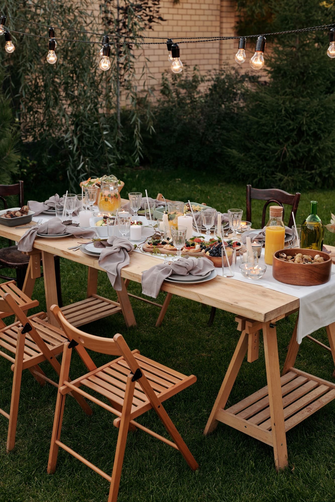 A long wooden table set for supper on the lawn, string lights in the trees above