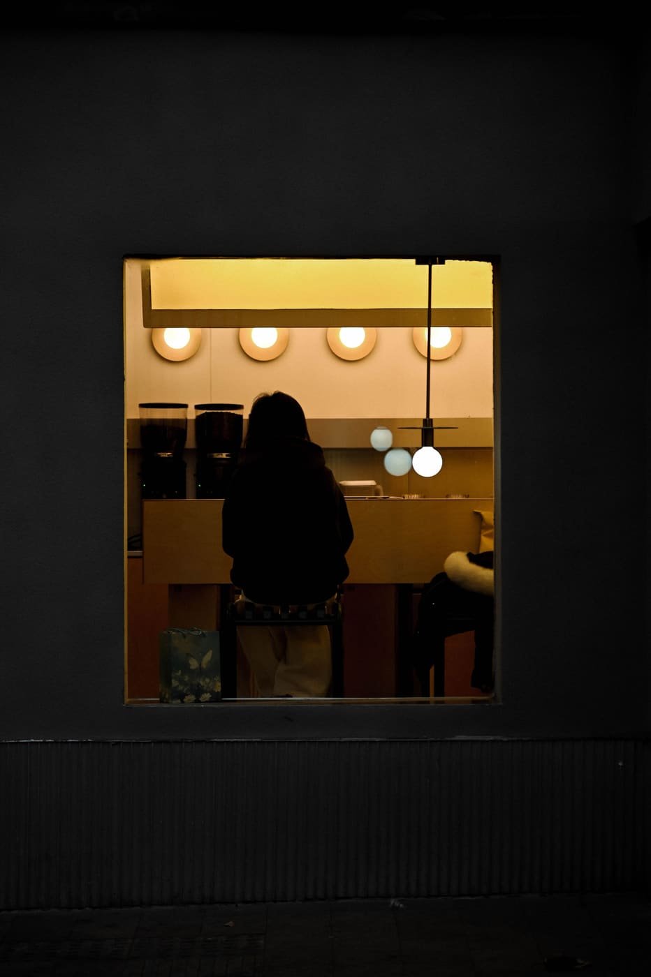 A person waiting at a warm lit counter under round pendant lamps, seen through a dark doorway at night