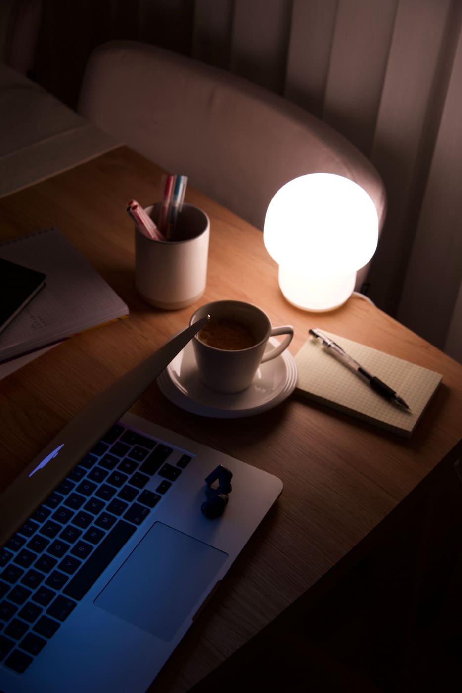 A lamp, a cup of coffee, a notebook and an open laptop on a wooden table at night