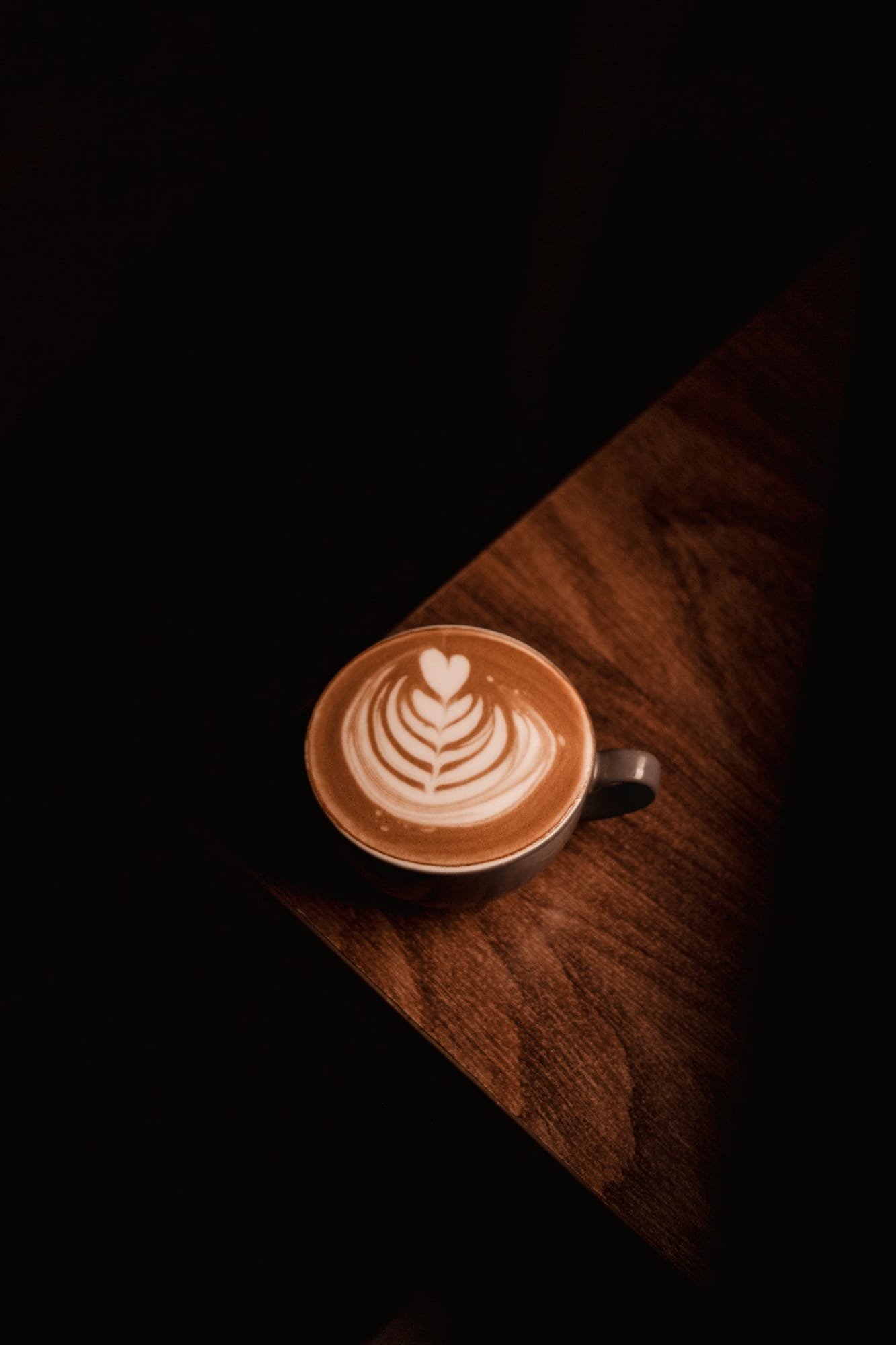 A latte with a leaf poured in the foam on the corner of a dark wood table, lit by one warm lamp at night
