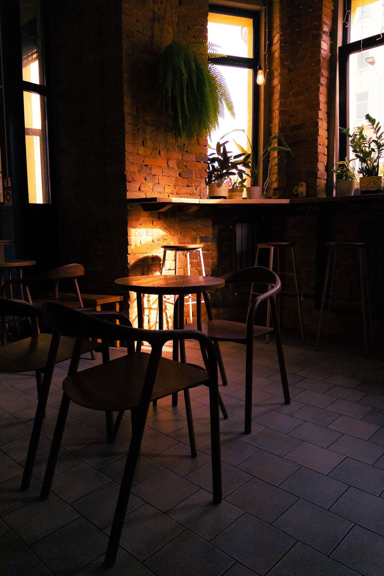 The coffee bar at night: brick walls, wooden stools and tables, and warm lamps over the counter