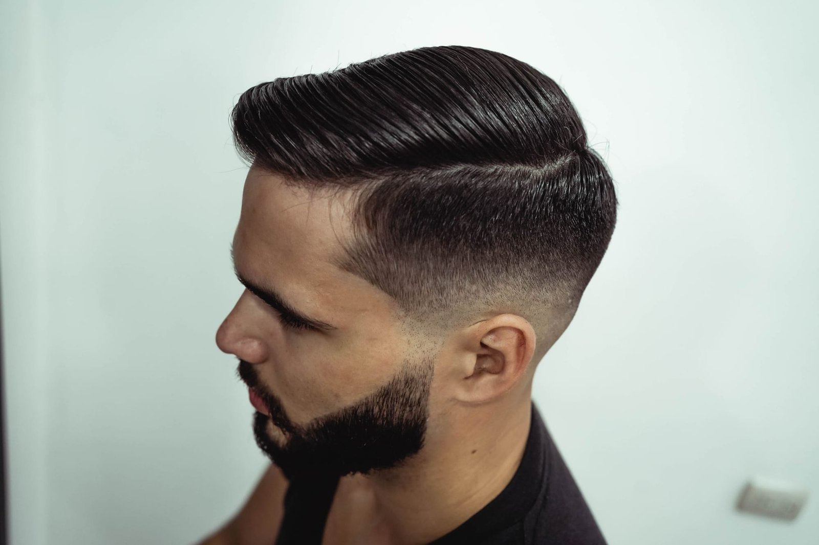 A fresh skin fade with a hard part and a trimmed beard, seen in profile against a pale wall