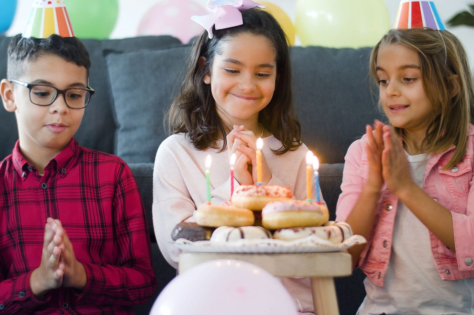 Three kids in party hats around a stack of donuts with lit birthday candles