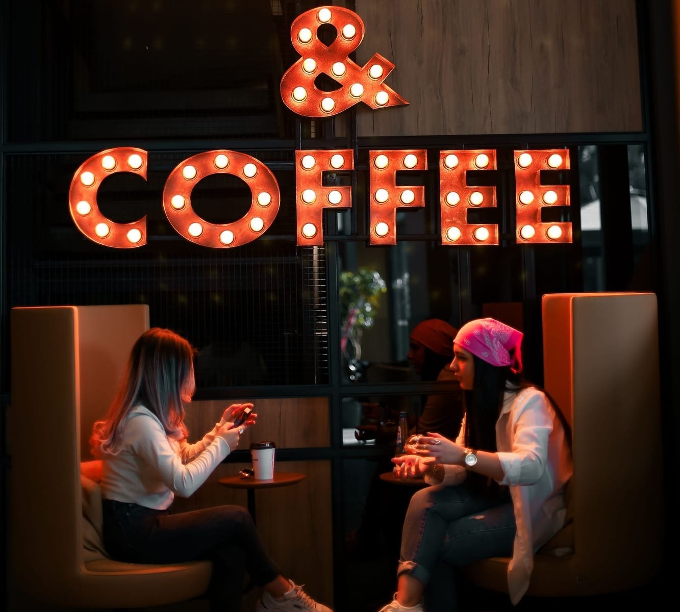 Two friends with coffee in a booth under a lit bulb sign