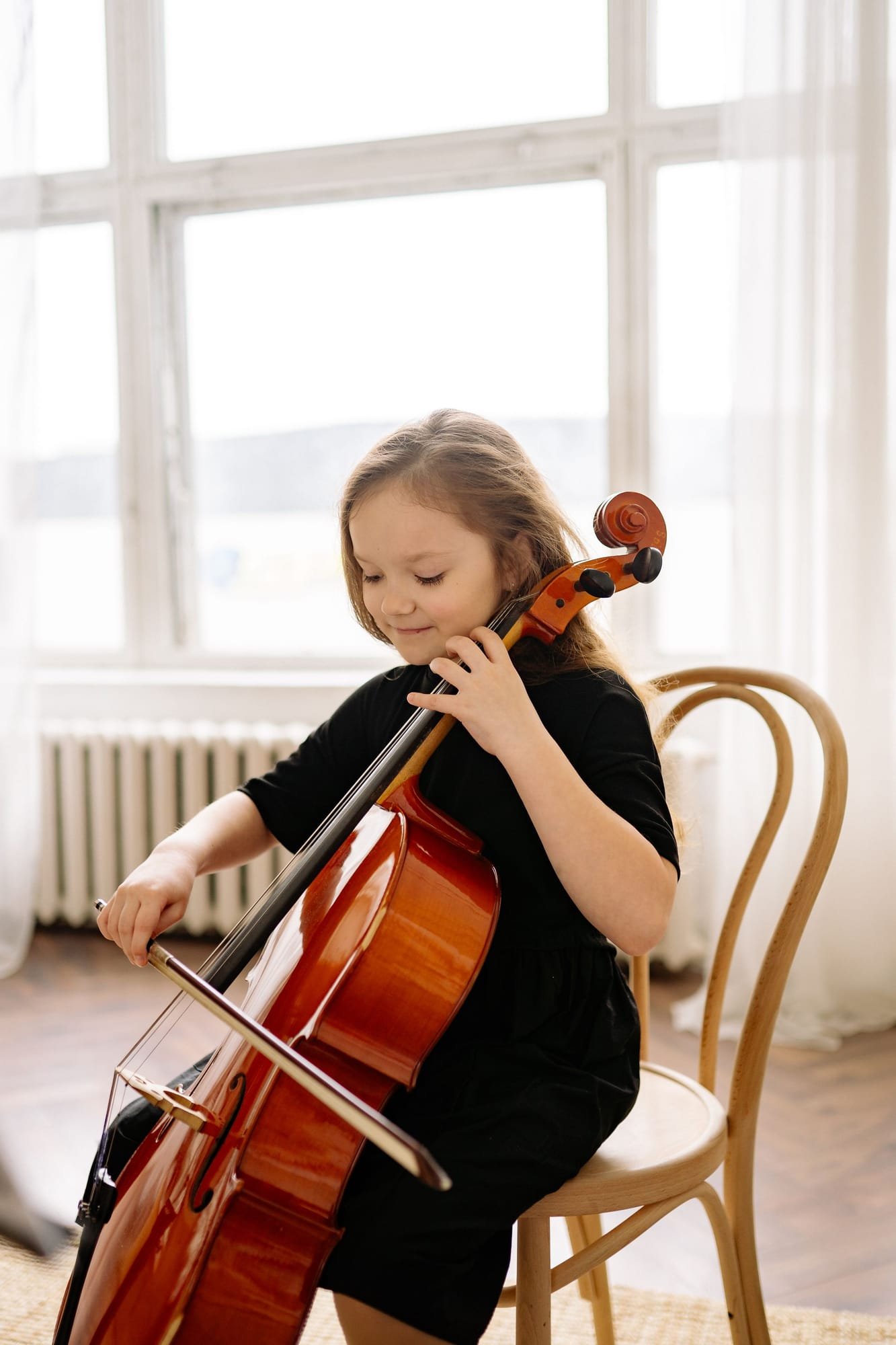 A young student with her eyes closed, playing the cello by a bright window