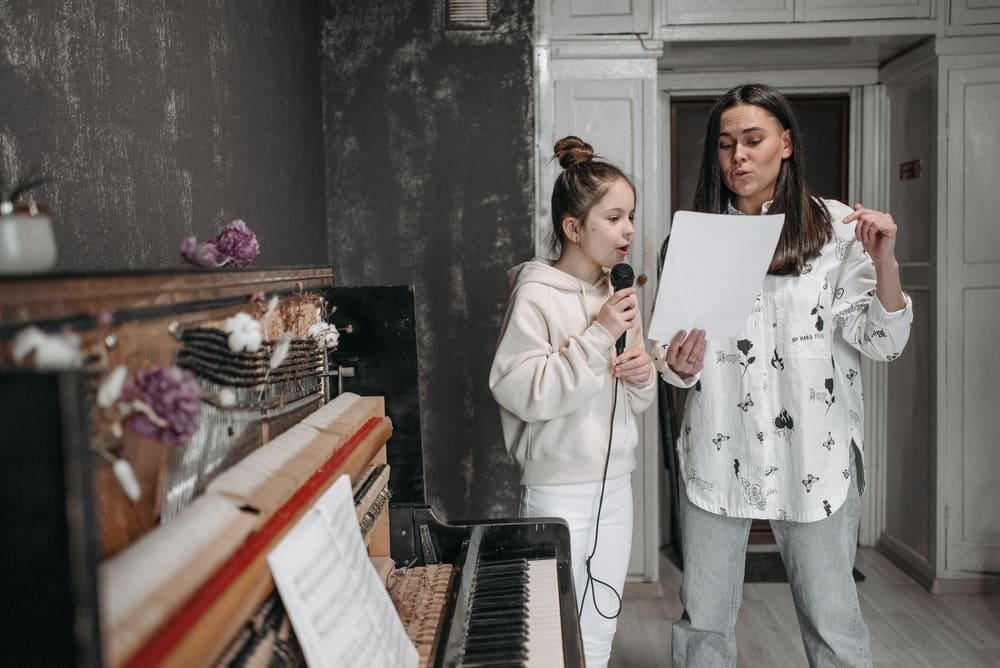 A voice teacher and a girl singing from sheet music beside an old upright piano