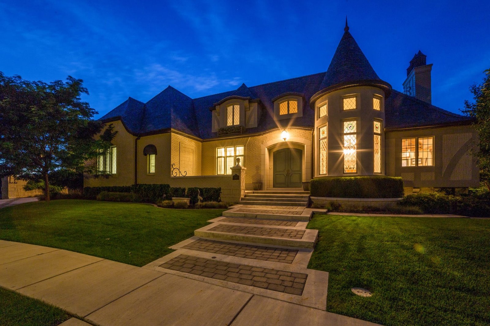 A stone and brick home with a round tower at dusk, every window lit, a wide front walk across the lawn